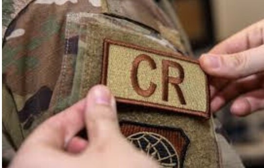 Hands pressing a tan embroidered patch with brown CR lettering onto a hook-and-loop panel on a camouflage uniform sleeve.