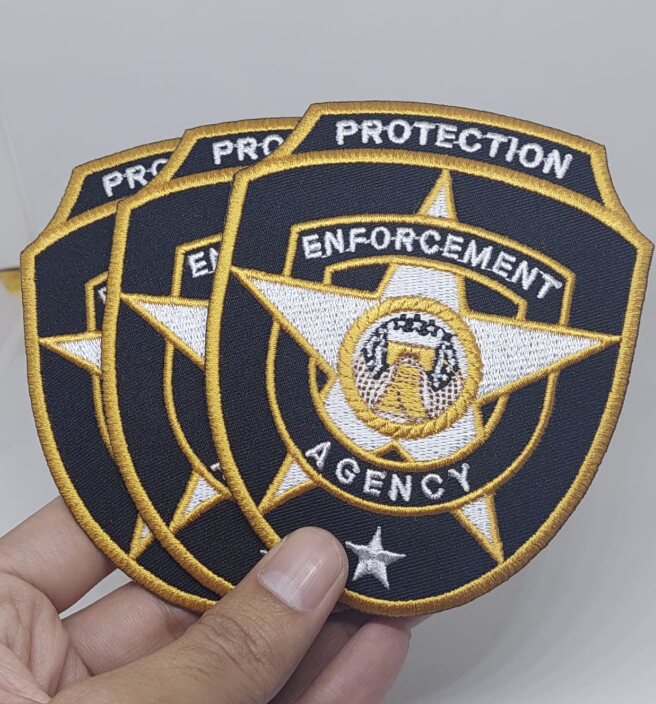 A hand holding three shield-shaped embroidered patches with black twill, white star, and gold merrowed borders and lettering.
