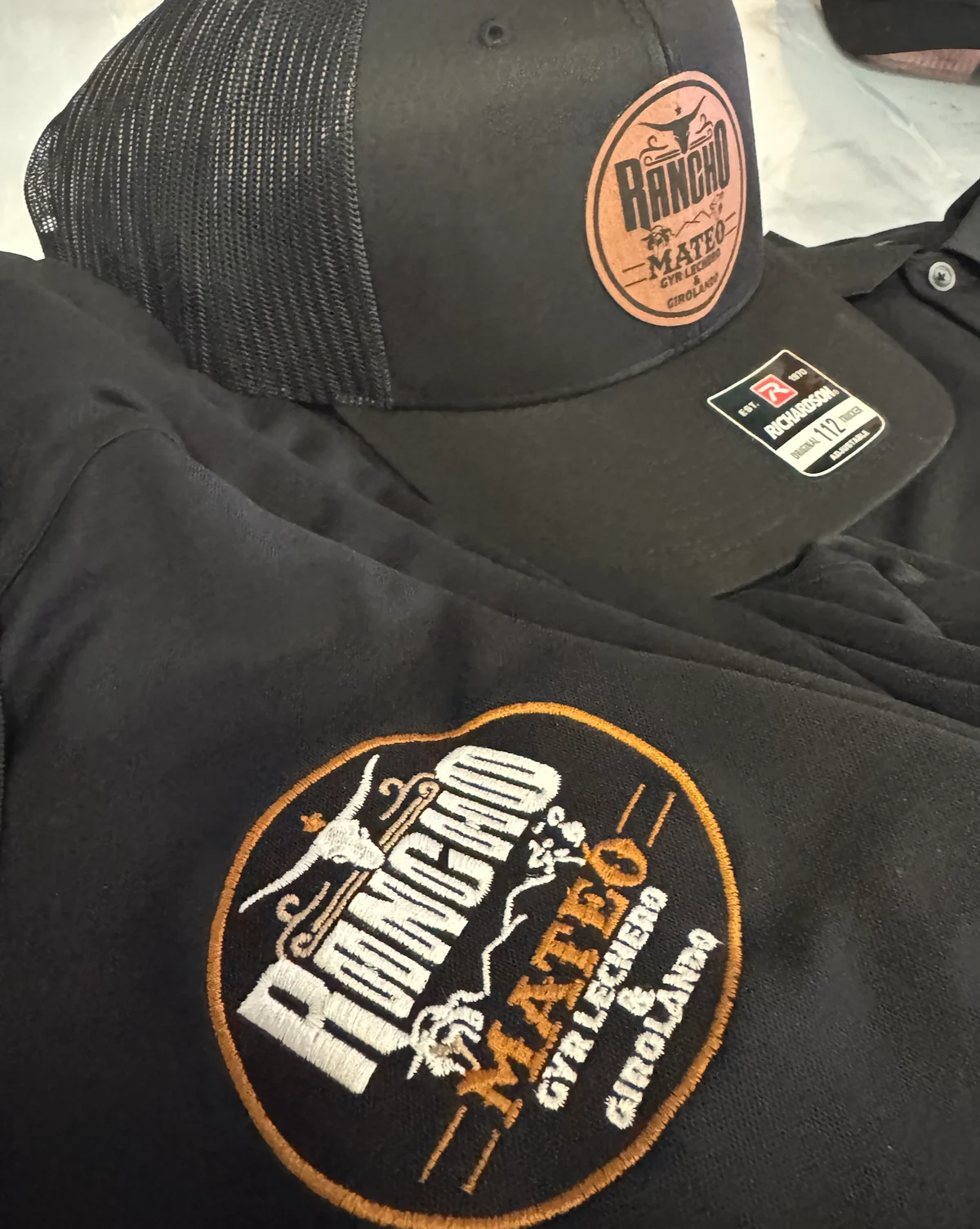 Black shirt with an orange and white Rancho Mateo embroidered logo, next to a matching leather-look patch cap